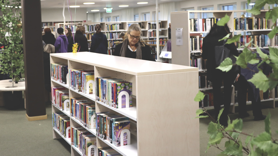 Hyllor med böcker, gröna växter och besökare i Vänersborgs bibliotek.