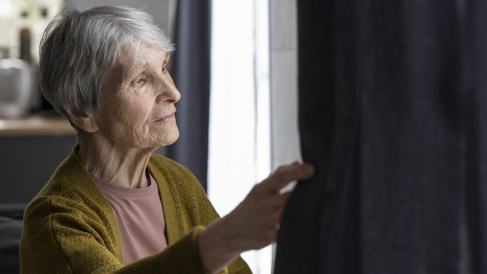 Äldre dam som tittar ut genom sitt fönster mellan mörka gardiner.