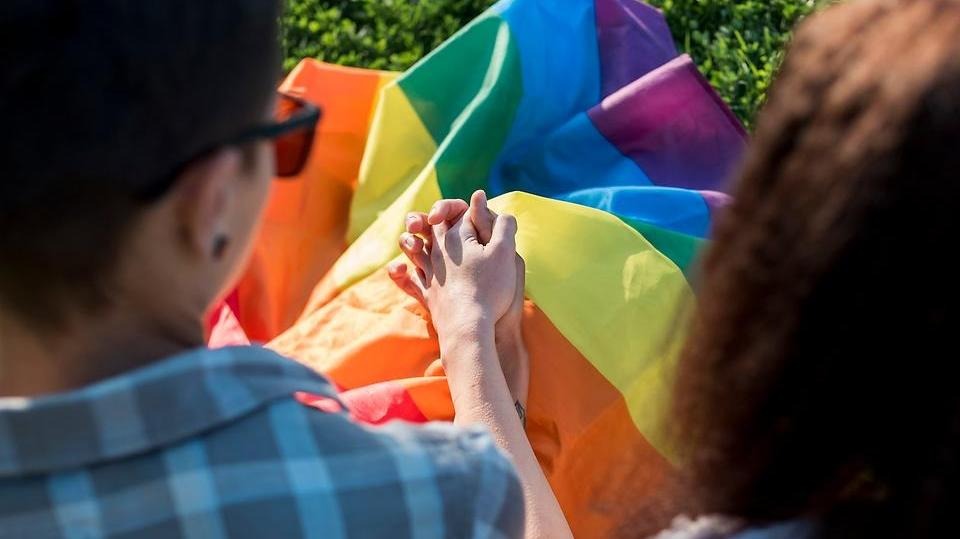 Två personer håller handen framför en regnbågsflagga