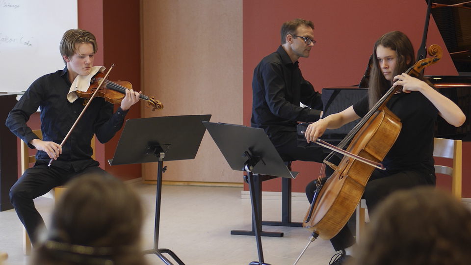 Två ungdomar som spelar stråkinstrument tillsammans med en man som spelar piano i bakgrunden.