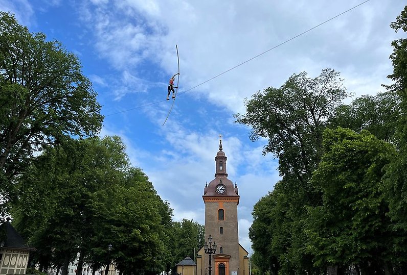 Verket Allting är möjligt, en lina mellan trädtoppar och Vänersborgs kyrktorn i bakgrunden. En skulptur föreställande en människa som balanserar på linan med röda och svarta kläder.