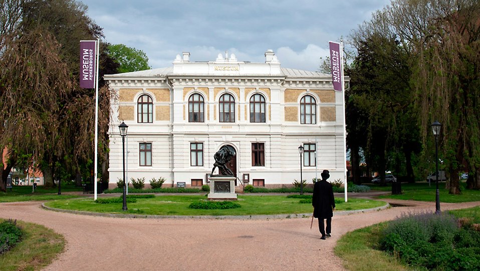 En person med hög hatt och svart rock går mot museets vita stenbyggnad.